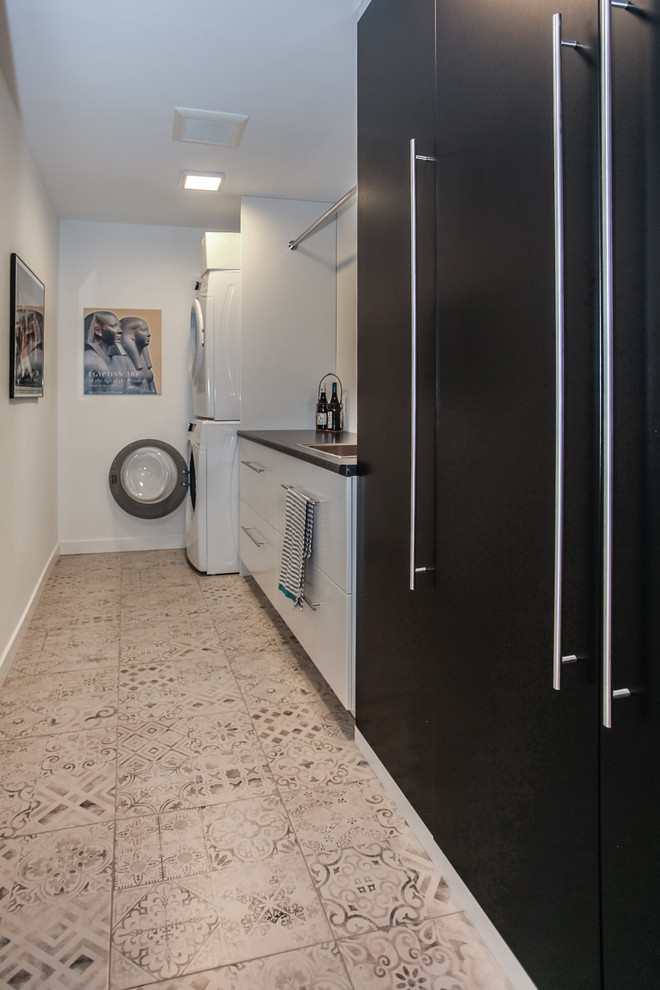 Design ideas for a mid-sized contemporary galley utility room in Toronto with a single-bowl sink, flat-panel cabinets, black cabinets, laminate benchtops, white walls, porcelain floors, a stacked washer and dryer and white floor.