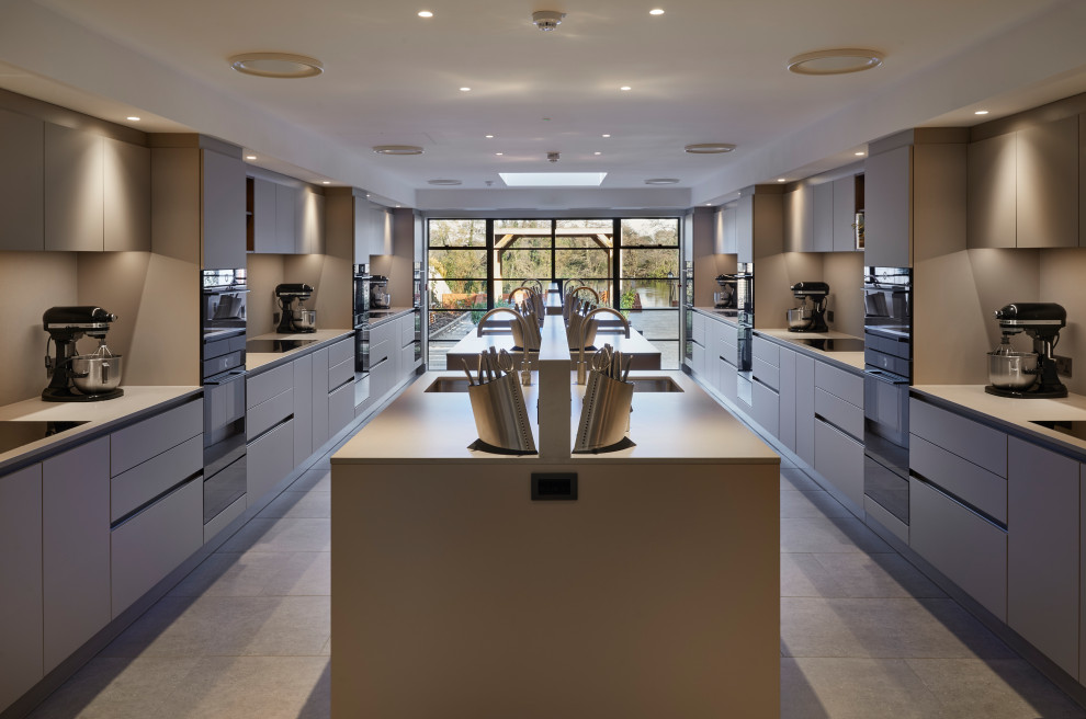 This is an example of an expansive contemporary grey and brown galley open plan kitchen in Berkshire with a single-bowl sink, flat-panel cabinets, beige cabinets, concrete worktops, grey splashback, engineered quartz splashback, black appliances, porcelain flooring, multiple islands, beige floors and grey worktops.