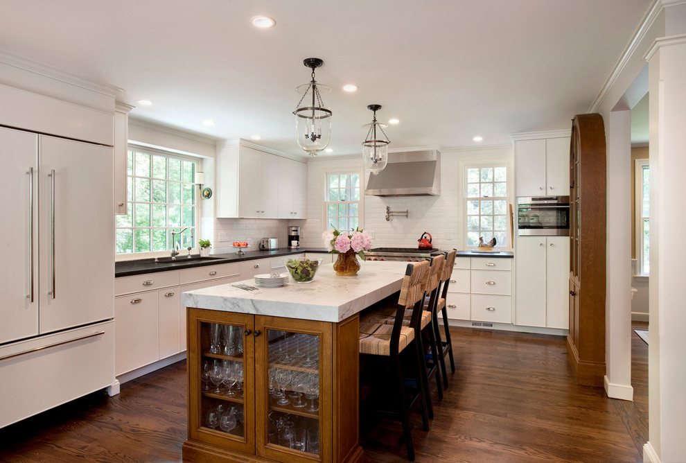 Transitional l-shaped eat-in kitchen in Boston with an undermount sink, flat-panel cabinets, white cabinets, white splashback, subway tile splashback, panelled appliances, medium hardwood floors, with island, brown floor and black benchtop.