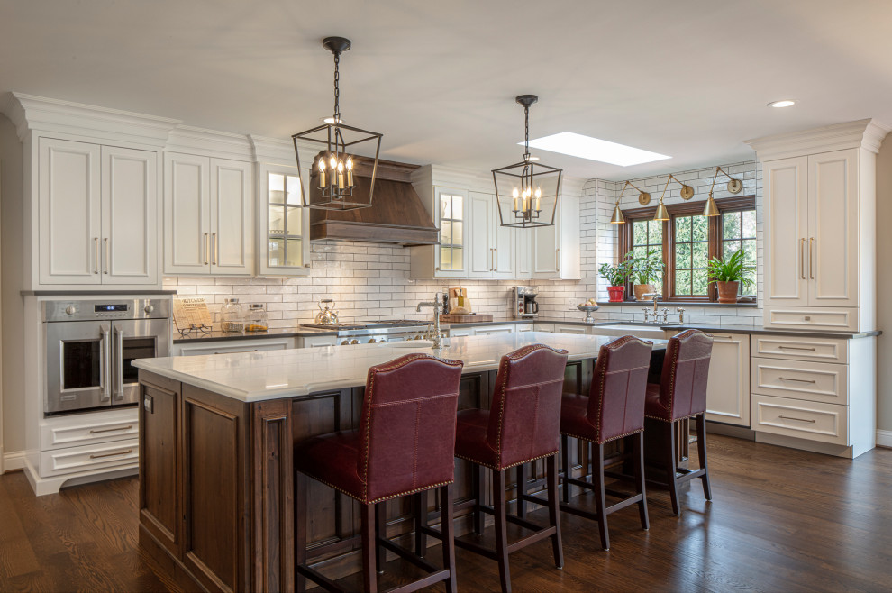 Traditional u-shaped eat-in kitchen in St Louis with a farmhouse sink, recessed-panel cabinets, white cabinets, white splashback, subway tile splashback, stainless steel appliances, medium hardwood floors, with island, brown floor and white benchtop.