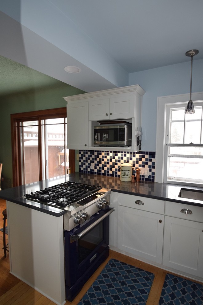 Example of a small transitional l-shaped light wood floor kitchen design with an undermount sink, shaker cabinets, white cabinets, quartz countertops, multicolored backsplash, ceramic backsplash, stainless steel appliances and a peninsula
