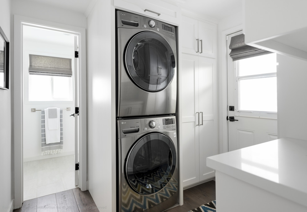 This is an example of a small transitional u-shaped utility room in Los Angeles with shaker cabinets, white cabinets, solid surface benchtops, white walls, medium hardwood floors and a stacked washer and dryer.