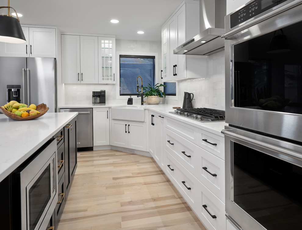 Mid-sized country l-shaped eat-in kitchen in Edmonton with a farmhouse sink, shaker cabinets, white cabinets, quartz benchtops, white splashback, ceramic splashback, stainless steel appliances, light hardwood floors, with island, brown floor and white benchtop.