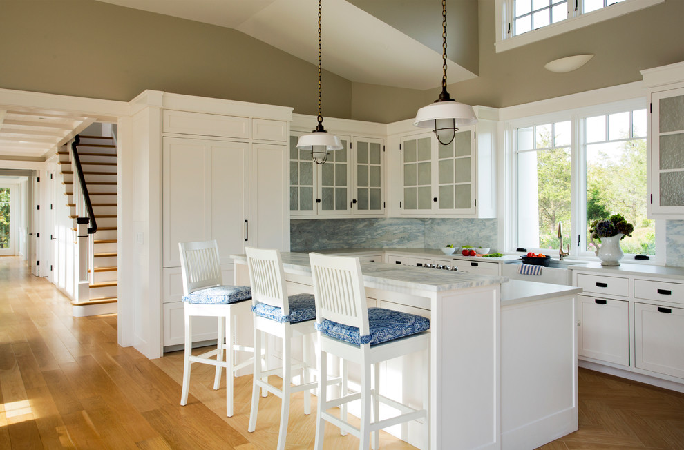 Photo of a mid-sized beach style l-shaped eat-in kitchen in Boston with a farmhouse sink, shaker cabinets, white cabinets, blue splashback, panelled appliances, medium hardwood floors, with island, marble benchtops, stone slab splashback, brown floor and grey benchtop.