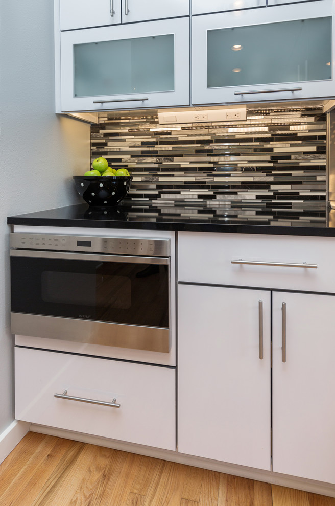 Large trendy u-shaped light wood floor open concept kitchen photo with an undermount sink, flat-panel cabinets, white cabinets, granite countertops, multicolored backsplash, mosaic tile backsplash, stainless steel appliances and an island