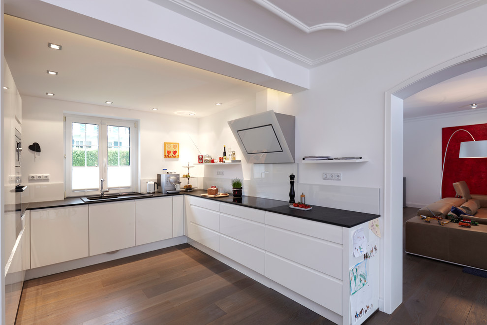 This is an example of a mid-sized contemporary u-shaped open plan kitchen in Essen with an integrated sink, flat-panel cabinets, white cabinets, white splashback, glass sheet splashback, dark hardwood floors and no island.