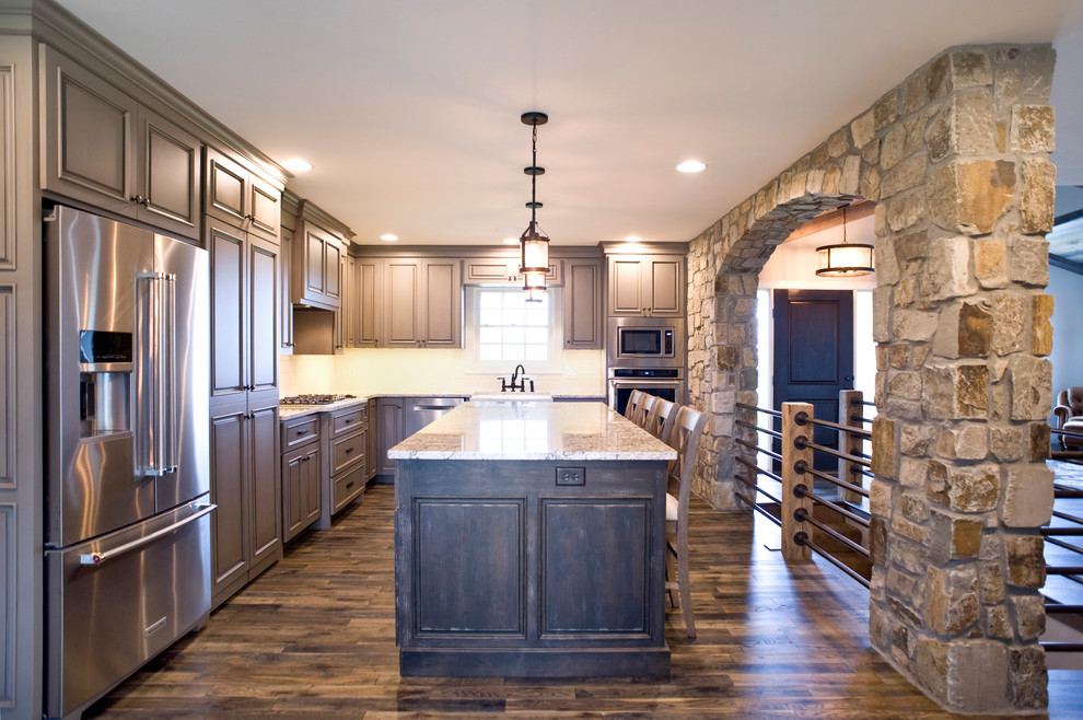Mid-sized transitional l-shaped medium tone wood floor and brown floor eat-in kitchen photo with a farmhouse sink, raised-panel cabinets, gray cabinets, marble countertops, white backsplash, subway tile backsplash, stainless steel appliances and an island