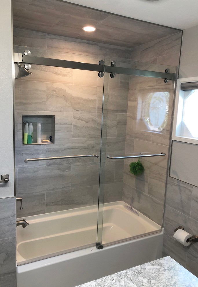 Inspiration for a mid-sized transitional 3/4 bathroom in Tampa with shaker cabinets, grey cabinets, an alcove tub, a shower/bathtub combo, a one-piece toilet, gray tile, porcelain tile, grey walls, porcelain floors, an undermount sink, granite benchtops, grey floor, a sliding shower screen and grey benchtops.
