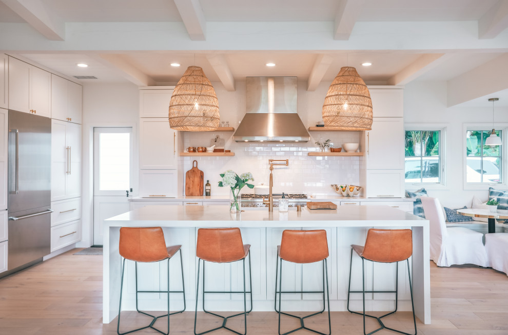 This is an example of a beach style l-shaped open plan kitchen in San Diego with a farmhouse sink, shaker cabinets, white cabinets, white splashback, subway tile splashback, light hardwood floors, with island, beige floor, white benchtop and exposed beam.