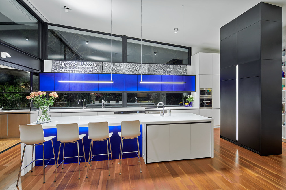 Inspiration for a large modern l-shaped open plan kitchen in Sydney with a double-bowl sink, flat-panel cabinets, blue cabinets, solid surface benchtops, panelled appliances, medium hardwood floors, with island and brown floor.