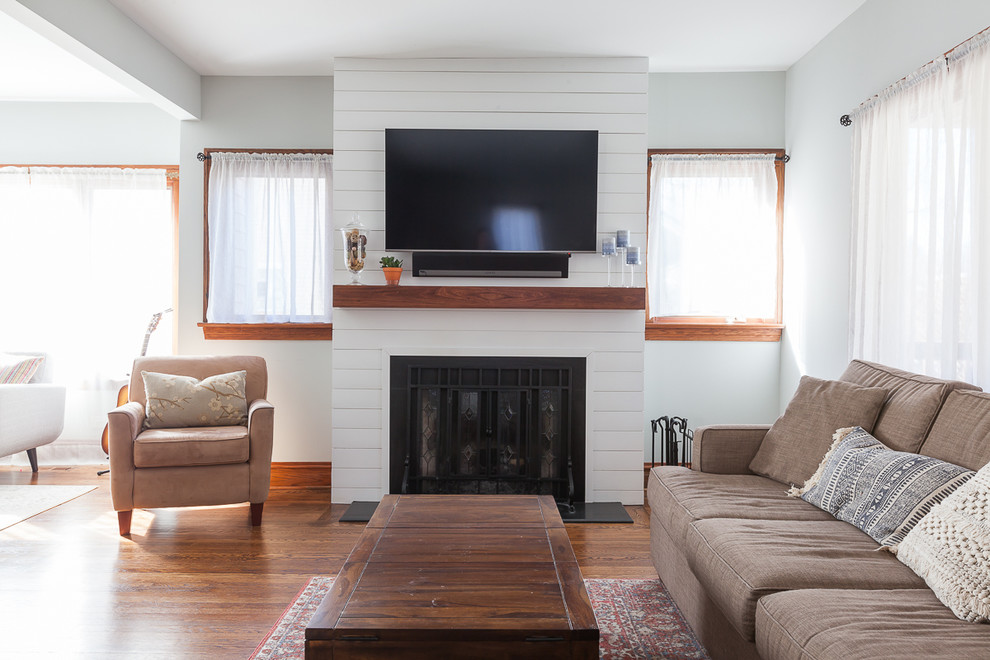 This is an example of a country open concept family room in Chicago with grey walls, medium hardwood floors, a standard fireplace, a wood fireplace surround, a wall-mounted tv and brown floor.