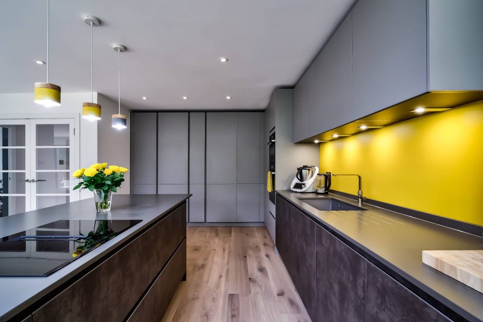 Photo of a large modern l-shaped open plan kitchen in London with flat-panel cabinets, grey cabinets, engineered stone countertops, yellow splashback, black appliances, an island and grey worktops.