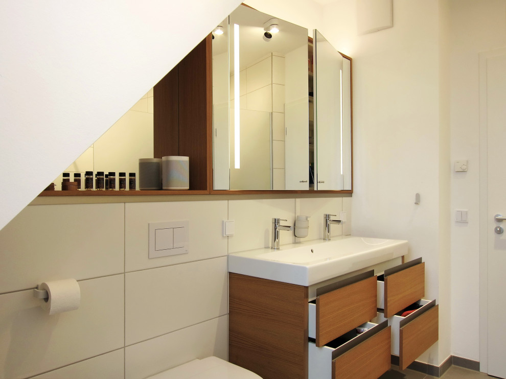 Пример оригинального дизайна: серо-белая ванная комната среднего размера со стиральной машиной в современном стиле с плоскими фасадами, светлыми деревянными фасадами, душем без бортиков, инсталляцией, белой плиткой, керамической плиткой, белыми стенами, полом из керамогранита, душевой кабиной, настольной раковиной, серым полом, открытым душем, тумбой под две раковины и подвесной тумбой