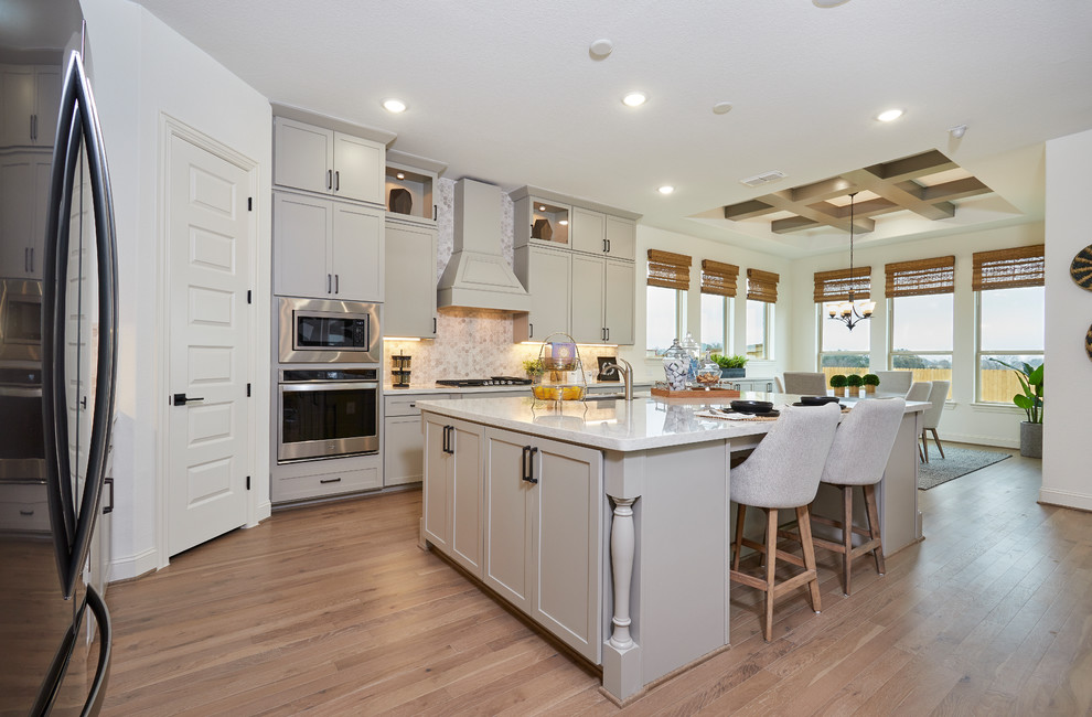 This is an example of a mid-sized traditional l-shaped eat-in kitchen in Other with an undermount sink, recessed-panel cabinets, grey cabinets, solid surface benchtops, beige splashback, stainless steel appliances, light hardwood floors, with island, beige floor and white benchtop.