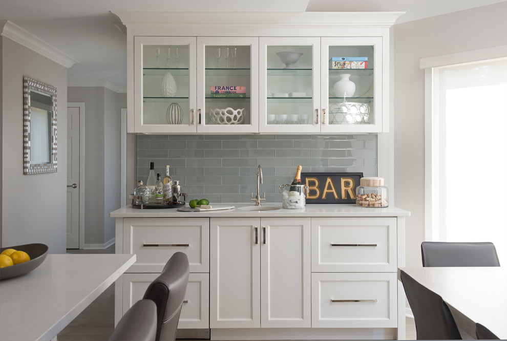 Small transitional single-wall wet bar in New York with an undermount sink, glass-front cabinets, white cabinets, quartz benchtops, grey splashback, glass tile splashback, porcelain floors, beige floor and white benchtop.