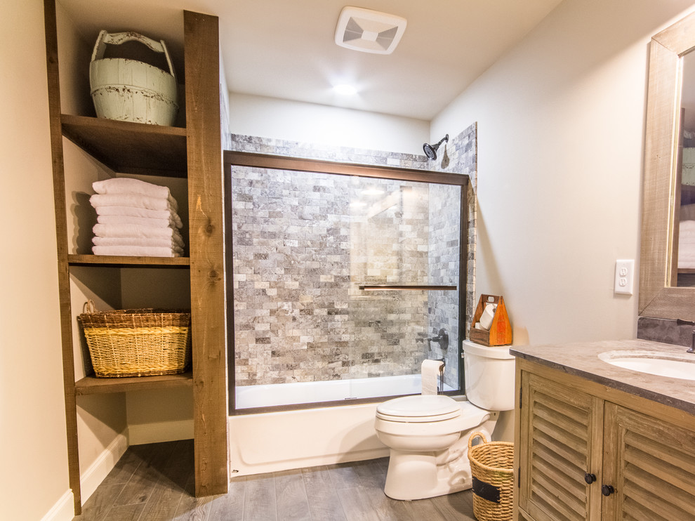 This is an example of a small country master bathroom in Atlanta with louvered cabinets, light wood cabinets, a drop-in tub, a shower/bathtub combo, a two-piece toilet, gray tile, stone tile, white walls, porcelain floors, an undermount sink, soapstone benchtops, grey floor, a sliding shower screen and grey benchtops.