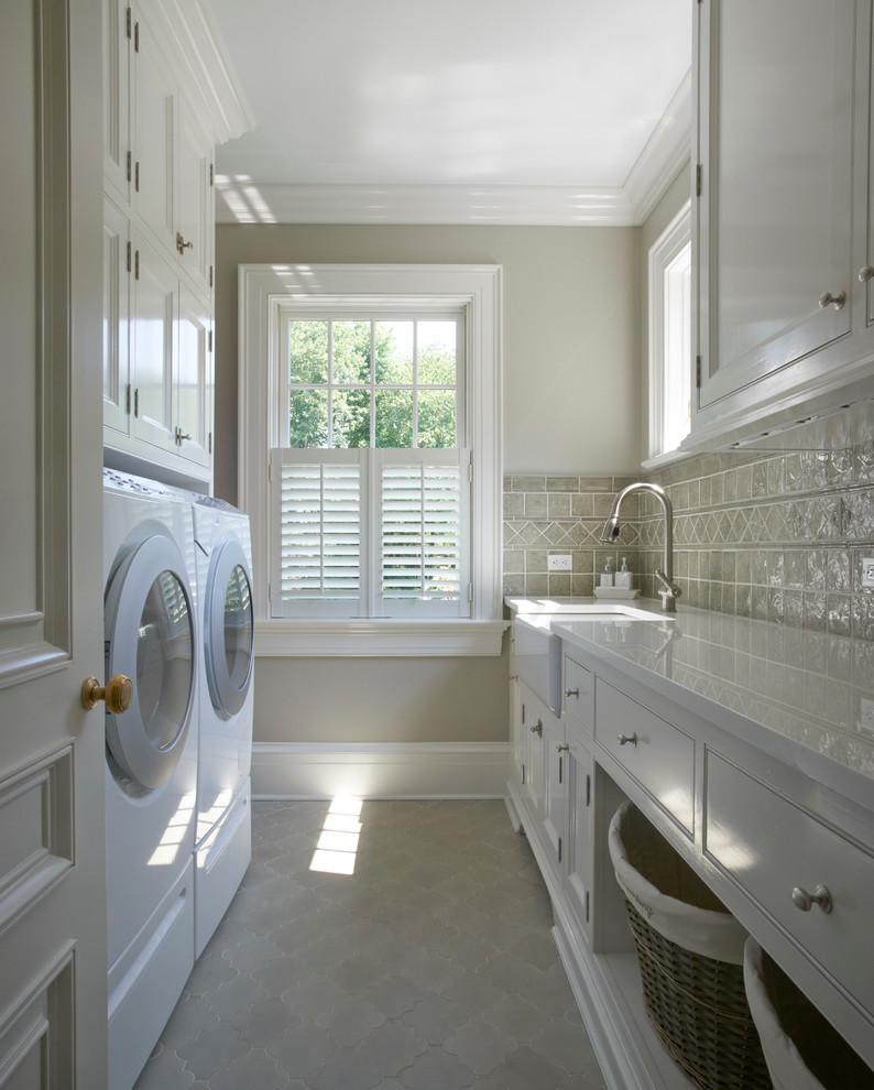This is an example of a mid-sized traditional galley dedicated laundry room in New York with white cabinets, a farmhouse sink, solid surface benchtops, ceramic floors, a side-by-side washer and dryer, grey floor, white benchtop, beaded inset cabinets and grey walls.