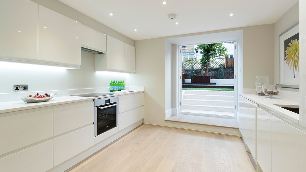 Photo of a contemporary galley separate kitchen in London with flat-panel cabinets, white cabinets and light hardwood floors.
