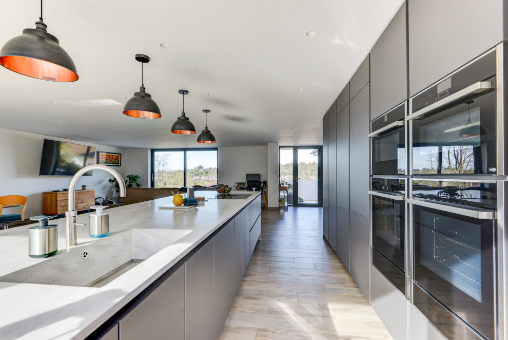 Bild på ett mycket stort funkis grå linjärt grått kök med öppen planlösning, med en integrerad diskho, släta luckor, grå skåp, bänkskiva i koppar, svarta vitvaror, ljust trägolv, en köksö och brunt golv