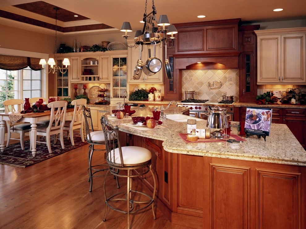 This is an example of a large traditional l-shaped open plan kitchen in Denver with a farmhouse sink, raised-panel cabinets, medium wood cabinets, granite benchtops, beige splashback, travertine splashback, stainless steel appliances, medium hardwood floors, with island, red floor and multi-coloured benchtop.