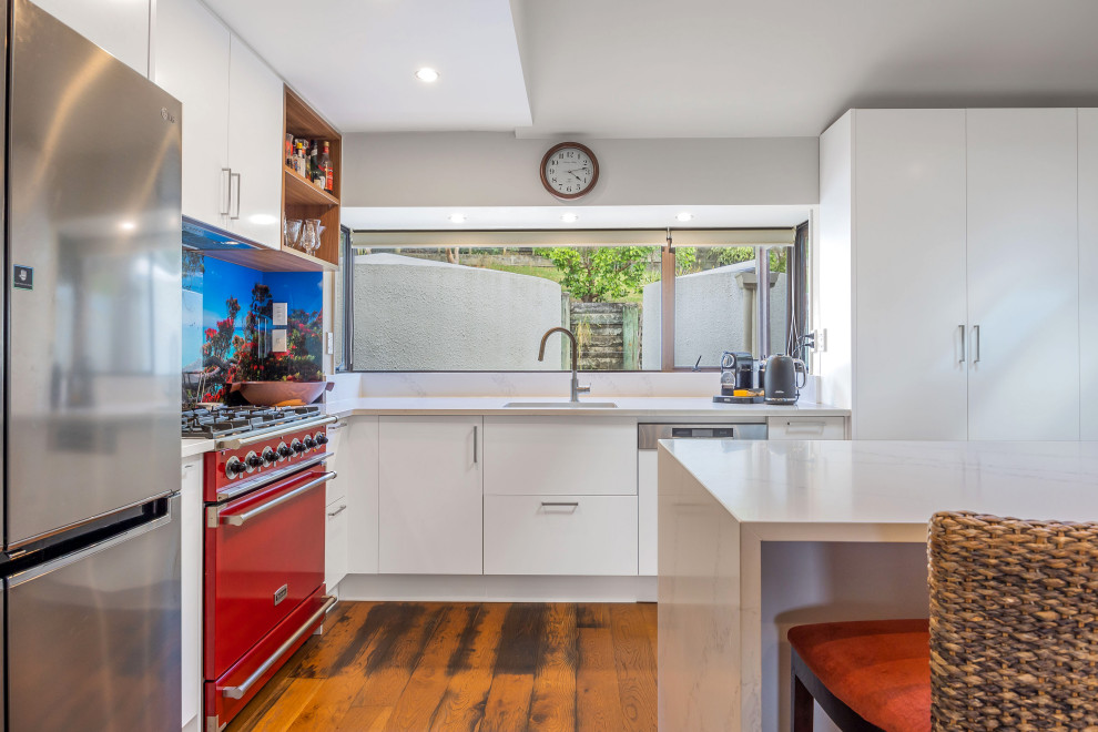 Small modern l-shaped eat-in kitchen in Auckland with an undermount sink, white cabinets, quartz benchtops, glass sheet splashback, coloured appliances, medium hardwood floors, with island and white benchtop.