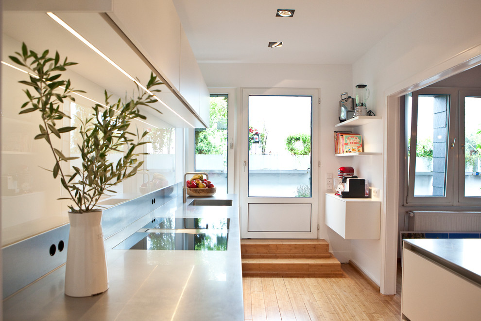 Design ideas for a mid-sized contemporary single-wall separate kitchen in Dusseldorf with an integrated sink, flat-panel cabinets, white cabinets, white splashback, light hardwood floors, no island, stainless steel benchtops and glass sheet splashback.