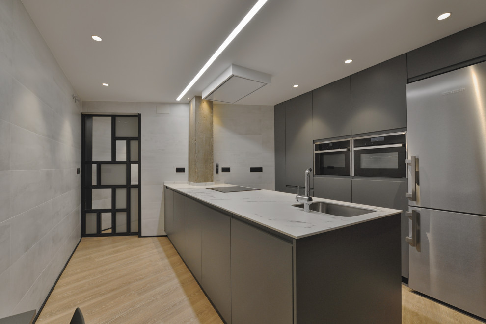Mid-sized industrial galley open plan kitchen in Madrid with an undermount sink, flat-panel cabinets, grey cabinets, limestone benchtops, stainless steel appliances, laminate floors, a peninsula, beige floor and white benchtop.