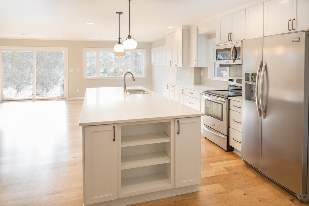 Mid-sized arts and crafts l-shaped open plan kitchen in Detroit with an undermount sink, shaker cabinets, white cabinets, solid surface benchtops, white splashback, subway tile splashback, stainless steel appliances, light hardwood floors and with island.