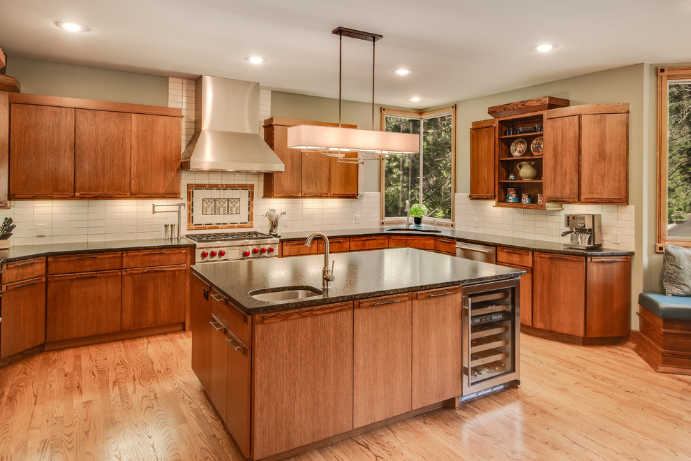 Large arts and crafts u-shaped kitchen in Grand Rapids with an undermount sink, flat-panel cabinets, medium wood cabinets, granite benchtops, white splashback, ceramic splashback, stainless steel appliances, light hardwood floors, with island, brown floor and black benchtop.