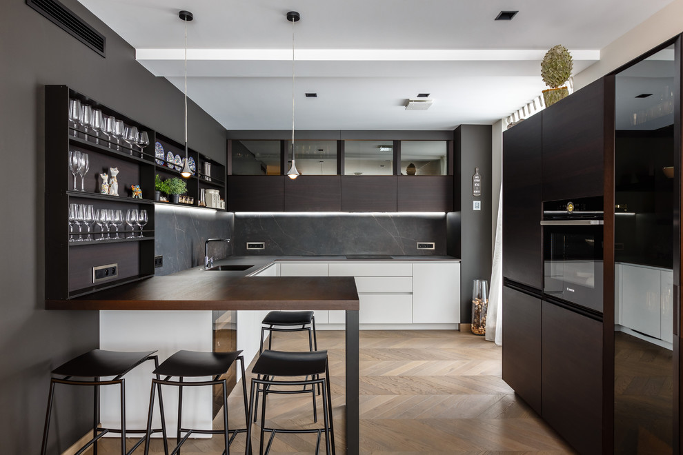 Photo of a contemporary u-shaped kitchen in Raleigh with an integrated sink, flat-panel cabinets, dark wood cabinets, black splashback, black appliances, light hardwood floors, beige floor and black benchtop.