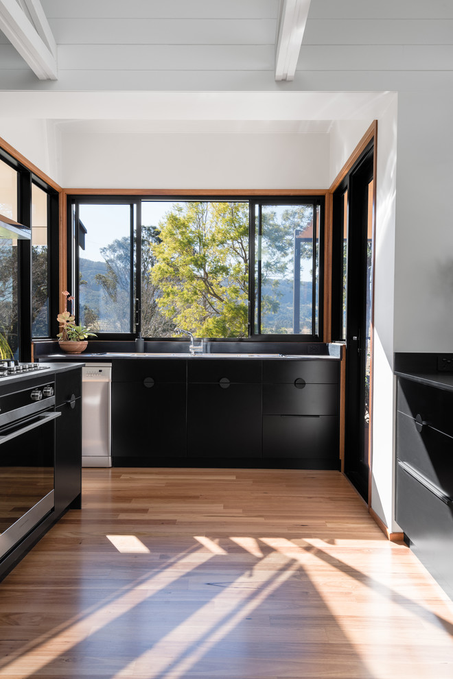 Large midcentury u-shaped eat-in kitchen in Other with a double-bowl sink, flat-panel cabinets, black cabinets, solid surface benchtops, window splashback, stainless steel appliances, medium hardwood floors, a peninsula and black benchtop.