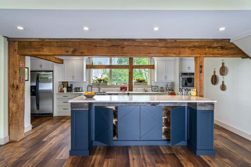 Inspiration for a medium sized rural galley open plan kitchen in Philadelphia with a belfast sink, shaker cabinets, blue cabinets, engineered stone countertops, white splashback, porcelain splashback, stainless steel appliances, medium hardwood flooring, an island, brown floors, white worktops and exposed beams.