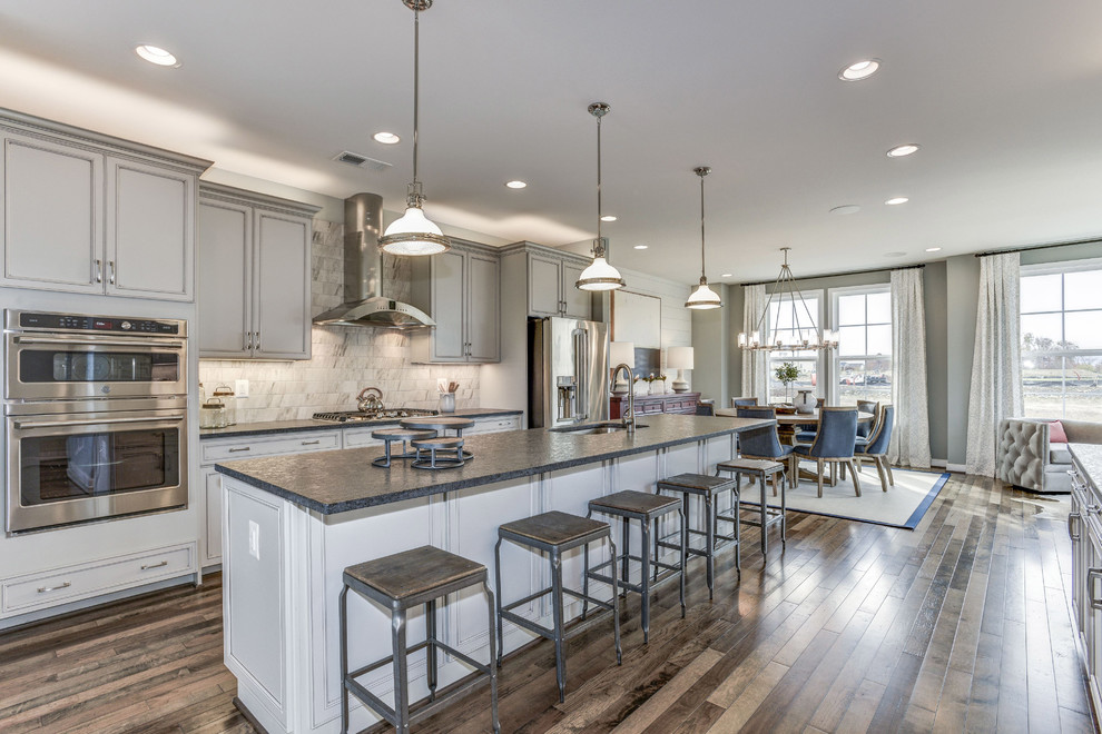 Photo of a country single-wall open plan kitchen in DC Metro with an undermount sink, recessed-panel cabinets, grey cabinets, grey splashback, stainless steel appliances, medium hardwood floors, with island, brown floor and black benchtop.
