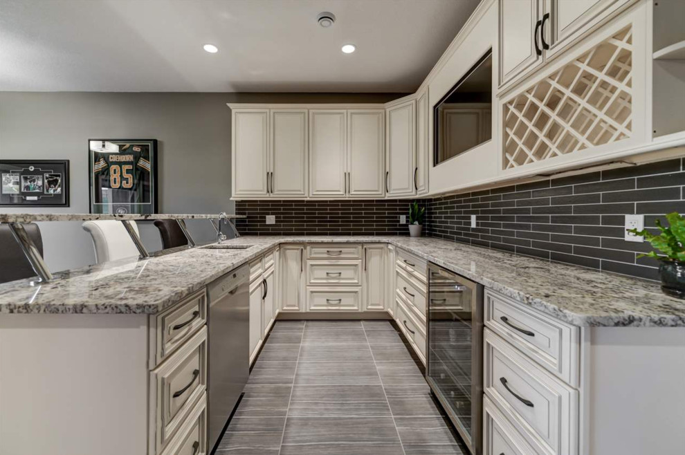 This is an example of an expansive arts and crafts u-shaped wet bar in Edmonton with a drop-in sink, beaded inset cabinets, beige cabinets, granite benchtops, brown splashback, glass tile splashback, porcelain floors and grey floor.