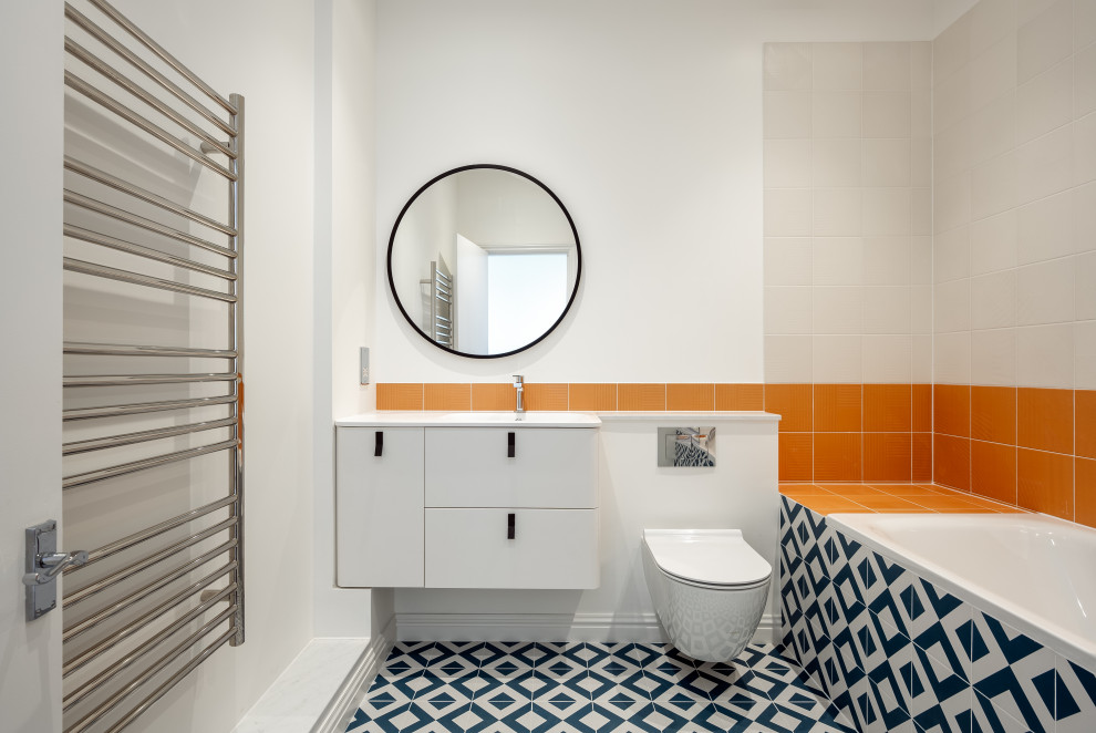 Mid-sized contemporary kids bathroom in London with flat-panel cabinets, white cabinets, a drop-in tub, a shower/bathtub combo, a wall-mount toilet, orange tile, ceramic tile, white walls, ceramic floors, a drop-in sink, marble benchtops, blue floor, a hinged shower door, white benchtops, a single vanity and a floating vanity.