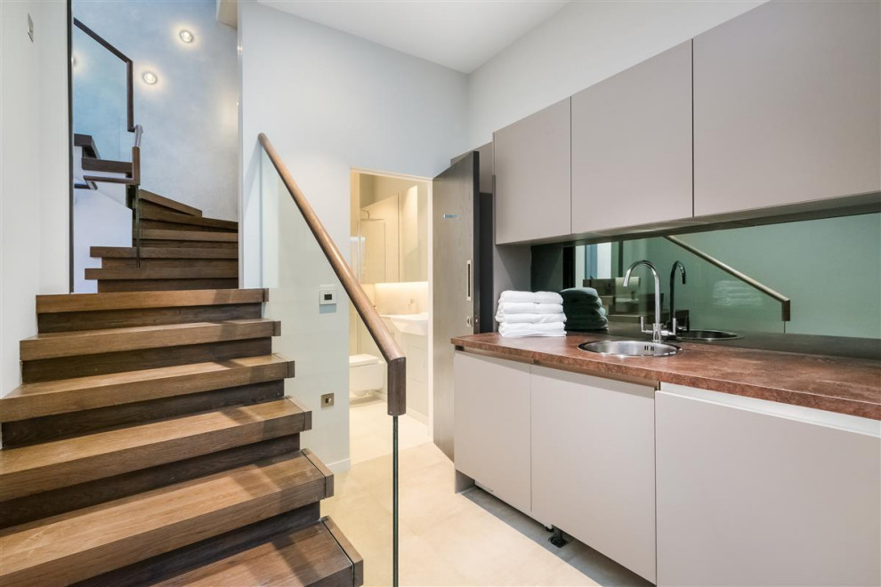Photo of a small contemporary single-wall utility room in London with a single-bowl sink, flat-panel cabinets, beige cabinets, wood benchtops, mirror splashback, porcelain floors and a concealed washer and dryer.