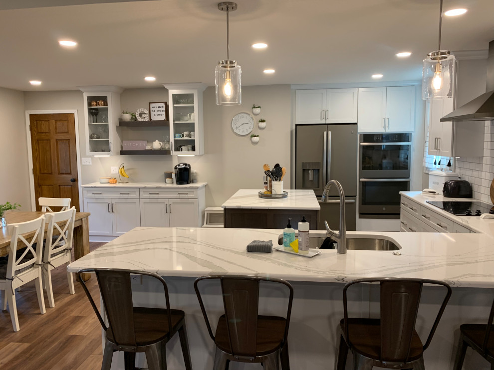 Open concept kitchen - large transitional u-shaped vinyl floor and brown floor open concept kitchen idea with an undermount sink, shaker cabinets, white cabinets, quartz countertops, white backsplash, ceramic backsplash, stainless steel appliances, an island and white countertops