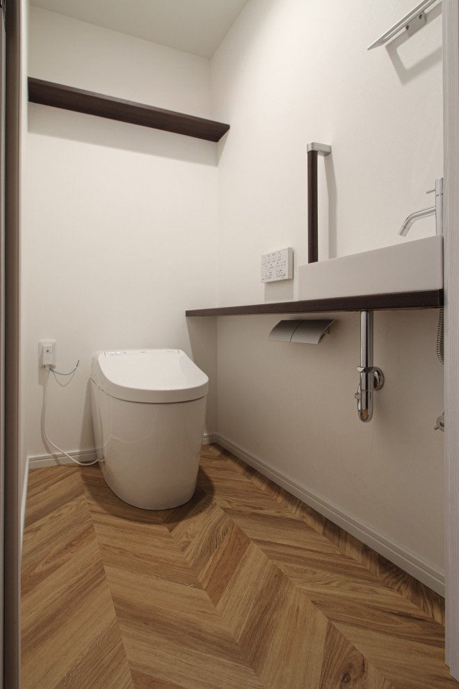 Bild på ett mellanstort minimalistiskt brun brunt toalett, med bruna skåp, en toalettstol med hel cisternkåpa, vita väggar, ljust trägolv, ett fristående handfat, träbänkskiva och brunt golv