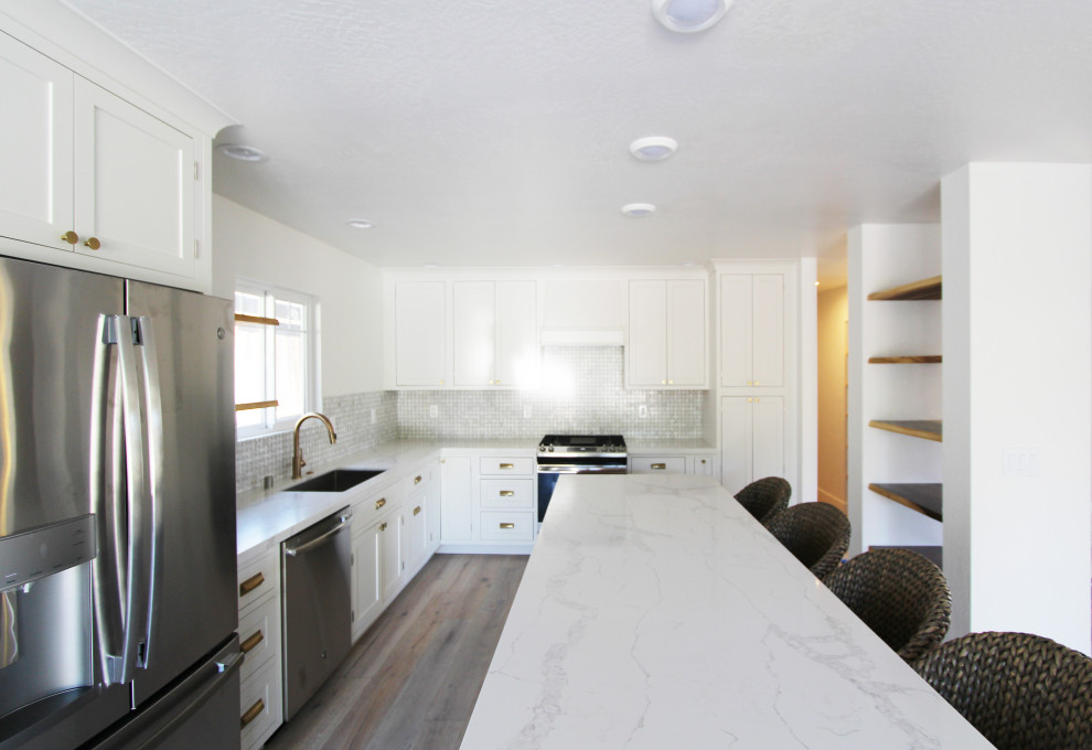 Example of a mid-sized l-shaped light wood floor and brown floor open concept kitchen design with an undermount sink, shaker cabinets, white cabinets, quartz countertops, gray backsplash, mosaic tile backsplash, stainless steel appliances, an island and white countertops