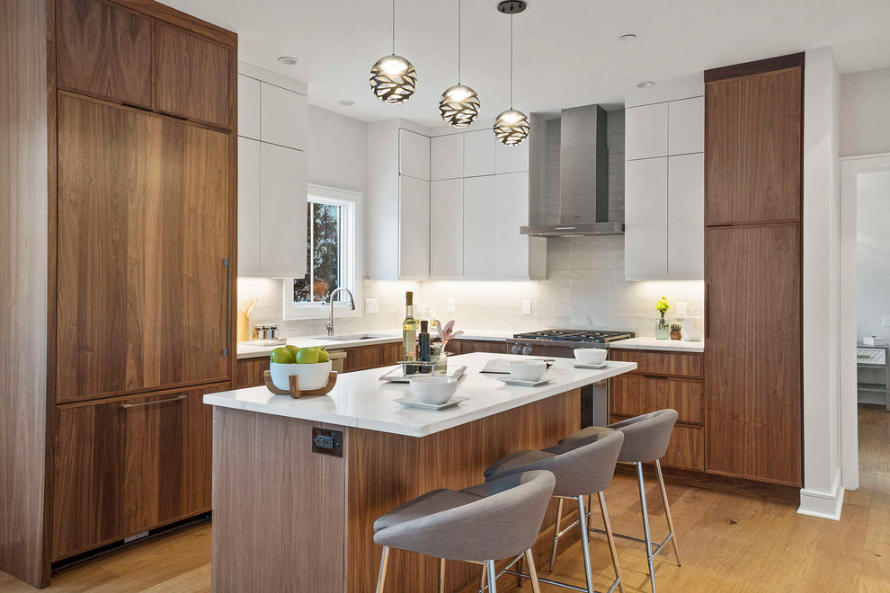 Mid-sized modern l-shaped eat-in kitchen in Boston with a drop-in sink, flat-panel cabinets, brown cabinets, quartzite benchtops, white splashback, ceramic splashback, stainless steel appliances, medium hardwood floors, with island, brown floor and white benchtop.