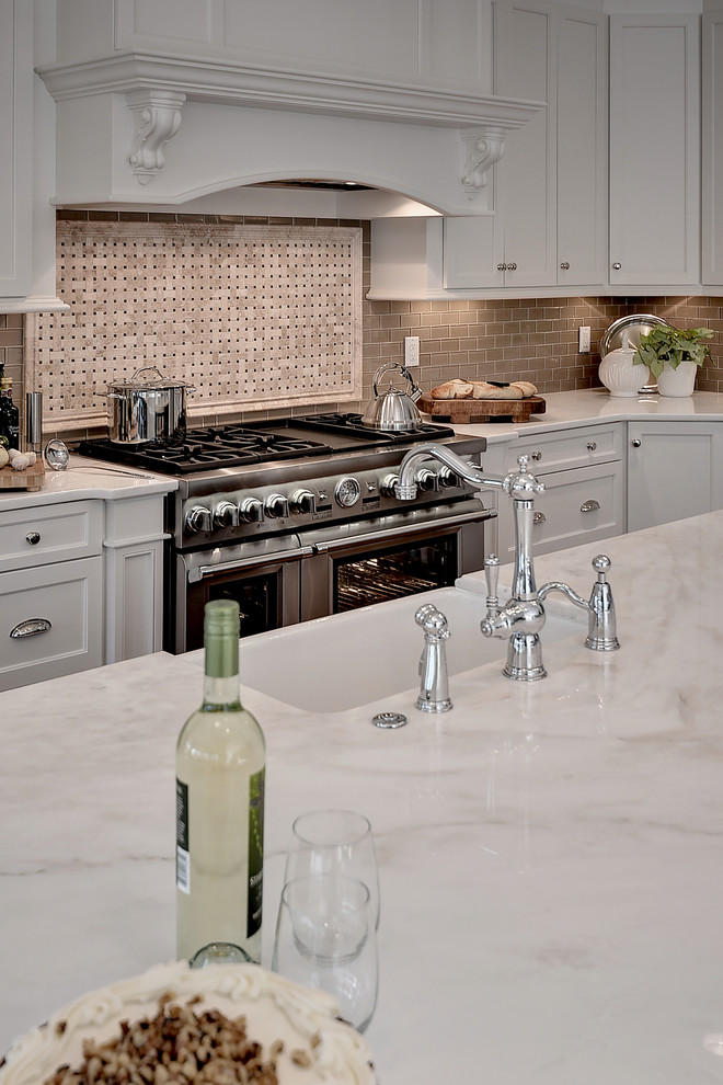 Huge transitional l-shaped medium tone wood floor and brown floor eat-in kitchen photo with a farmhouse sink, shaker cabinets, white cabinets, marble countertops, beige backsplash, glass tile backsplash, stainless steel appliances and an island