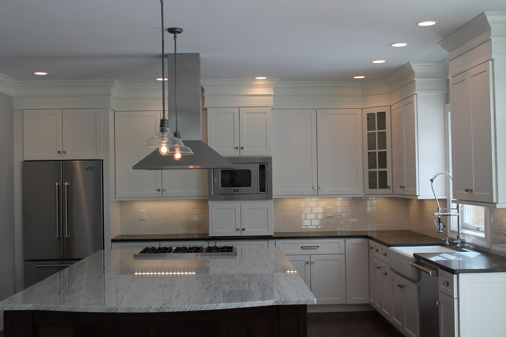This is an example of a contemporary l-shaped kitchen in New York with a farmhouse sink, shaker cabinets, white cabinets, granite benchtops, white splashback, subway tile splashback, stainless steel appliances, dark hardwood floors and with island.