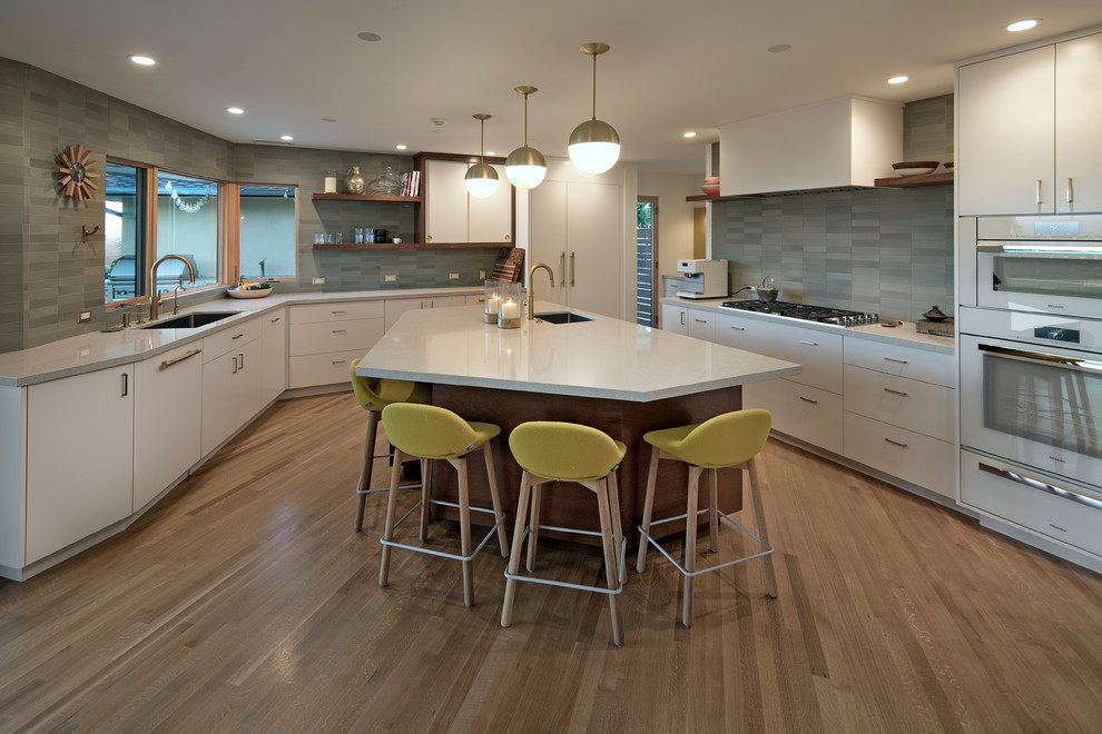 This is an example of a midcentury u-shaped kitchen in Santa Barbara with an undermount sink, flat-panel cabinets, white cabinets, grey splashback, white appliances, medium hardwood floors, with island, brown floor and white benchtop.