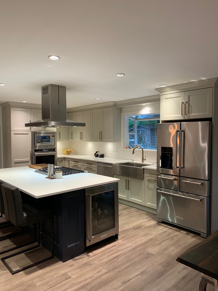 Large arts and crafts l-shaped open plan kitchen in Vancouver with a farmhouse sink, shaker cabinets, beige cabinets, quartz benchtops, white splashback, ceramic splashback, stainless steel appliances, laminate floors, with island, grey floor and white benchtop.