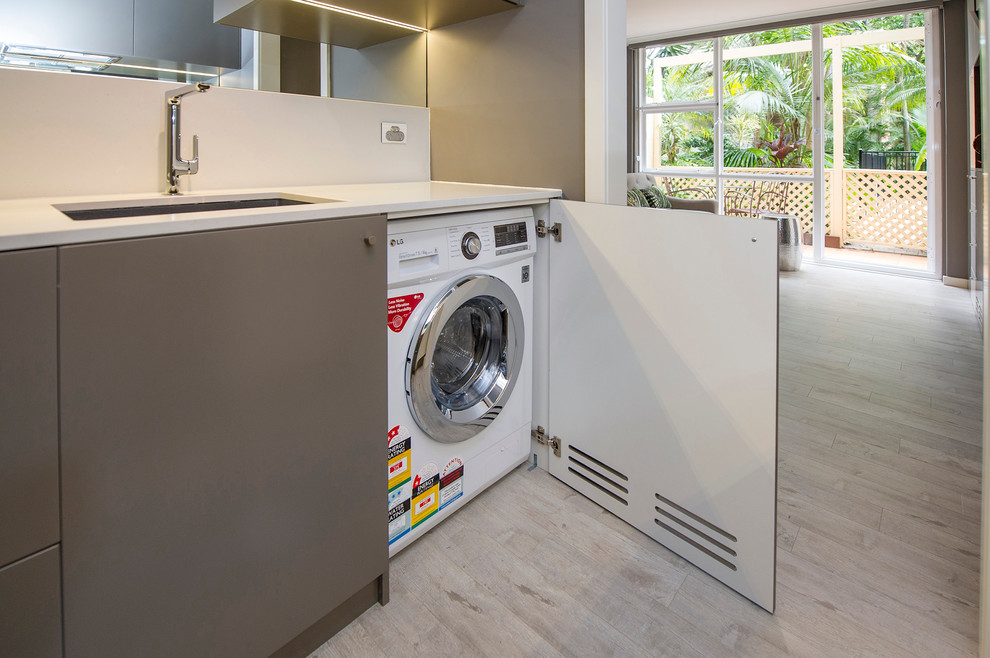 This is an example of a small contemporary galley utility room in Sydney with a single-bowl sink, flat-panel cabinets, solid surface benchtops, light hardwood floors and a concealed washer and dryer.