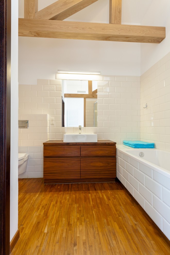 This is an example of a mid-sized midcentury 3/4 bathroom in Los Angeles with a drop-in sink, flat-panel cabinets, medium wood cabinets, wood benchtops, a drop-in tub, a wall-mount toilet, white tile, matchstick tile, white walls and light hardwood floors.
