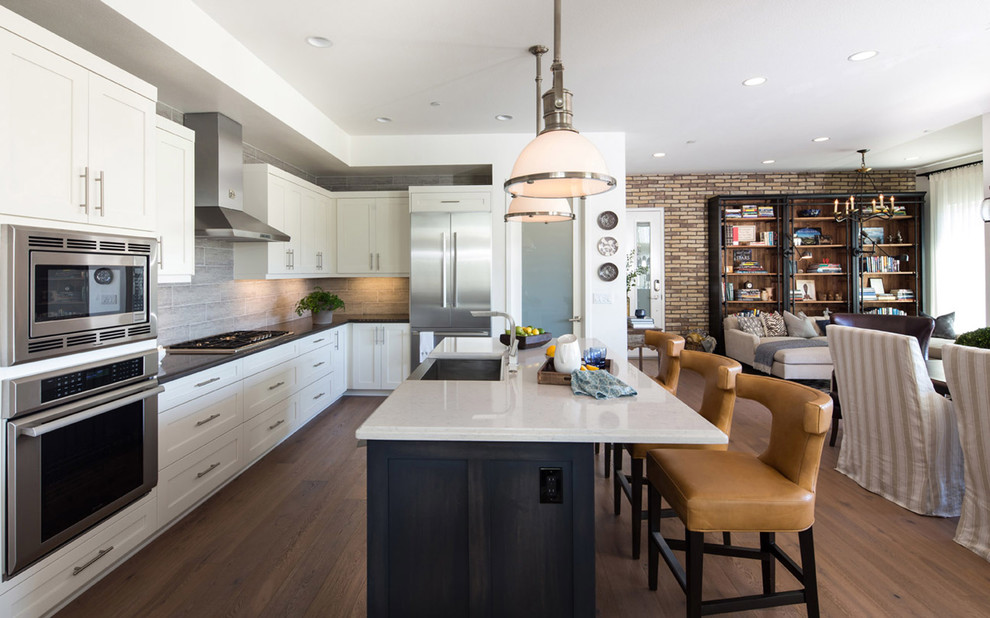 Photo of a mid-sized country l-shaped eat-in kitchen in Los Angeles with a farmhouse sink, shaker cabinets, white cabinets, quartz benchtops, grey splashback, porcelain splashback, white appliances, medium hardwood floors and with island.