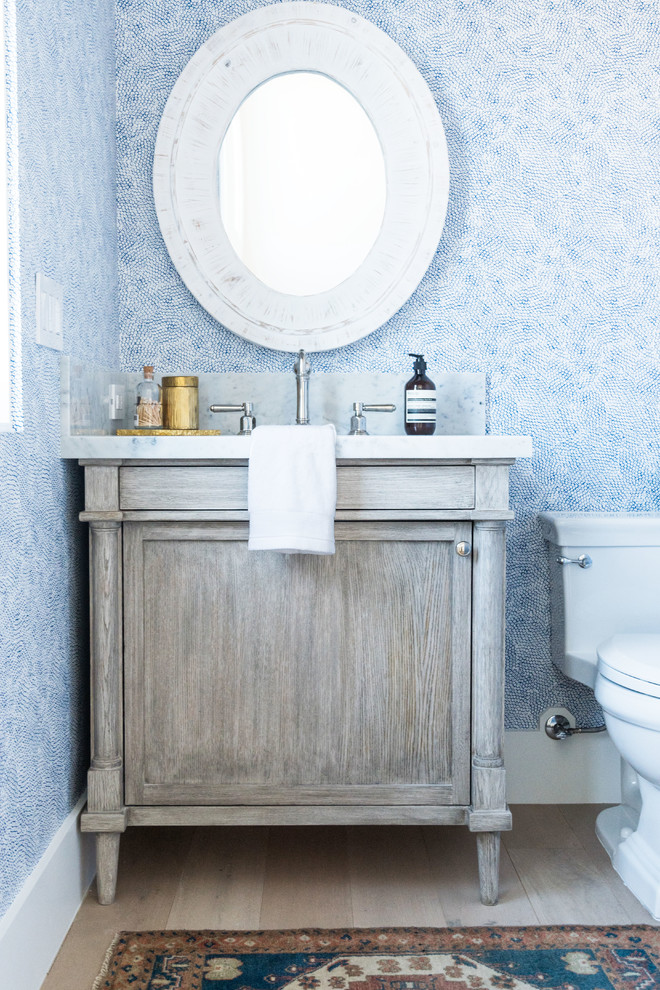 Beach style powder room in Orange County with furniture-like cabinets, grey cabinets, a one-piece toilet, blue walls, light hardwood floors, beige floor, white benchtops and marble benchtops.