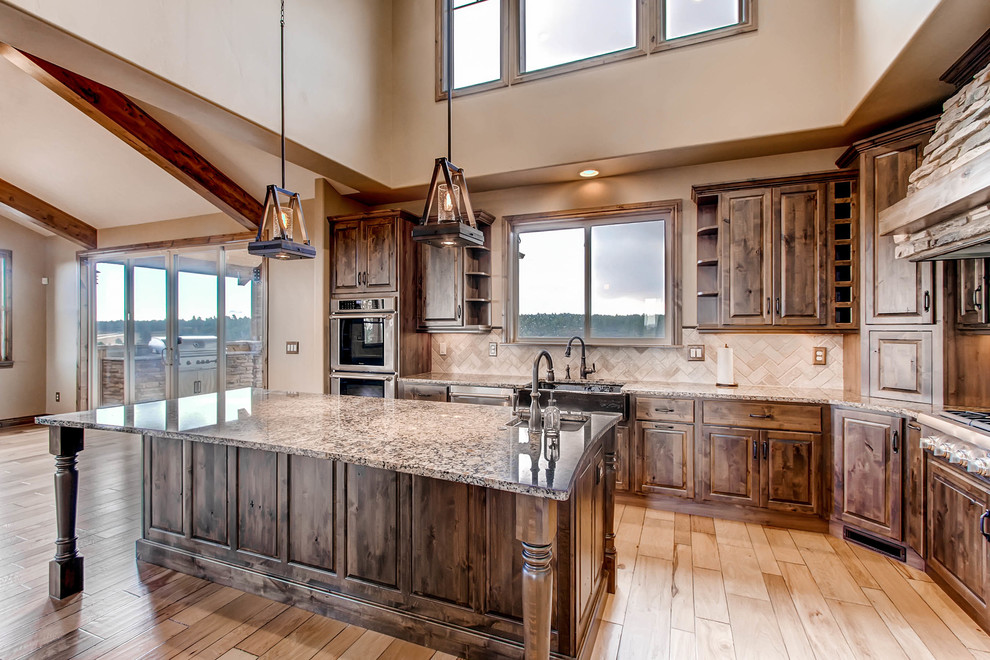 This is an example of a large country u-shaped kitchen in Denver with a farmhouse sink, raised-panel cabinets, dark wood cabinets, granite benchtops, beige splashback, stone tile splashback, stainless steel appliances, light hardwood floors, with island, brown floor and multi-coloured benchtop.