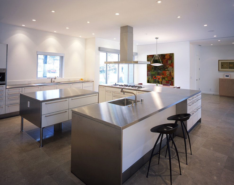 This is an example of a large modern l-shaped separate kitchen in Portland with an undermount sink, flat-panel cabinets, white cabinets, stainless steel benchtops, white splashback, stainless steel appliances, limestone floors, multiple islands and timber splashback.